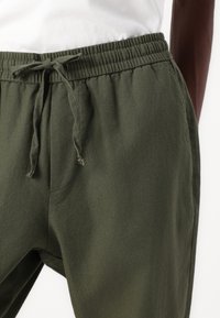 Joggebukser i olivengrønn har elastisk midjebånd med snor. Stoffet virker lett med en glatt tekstur og avslappet passform.