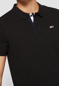 Svart polotröja med krage, som har en placket i vitt och marinblått. Litet märke på bröstet, textil med struktur. Kort ärm.