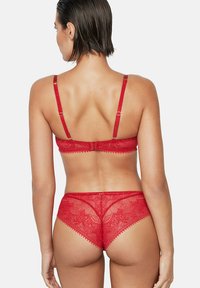 Vrouw met kort donker haar die een rood kanten beha en bijpassende kanten slip draagt, gezien van achter tegen een effen lichte achtergrond.