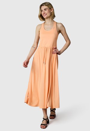 Femme souriante portant une robe maxi sans manches de couleur pêche avec taille à cordon et sandales noires à talons et brides, debout devant un fond clair.