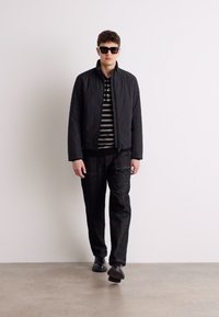 Jachetă neagră matlasată peste o cămașă cu dungi, asociată cu pantaloni închiși la culoare și pantofi negri. Modelul poartă ochelari de soare și stă în fața unui fundal simplu.
