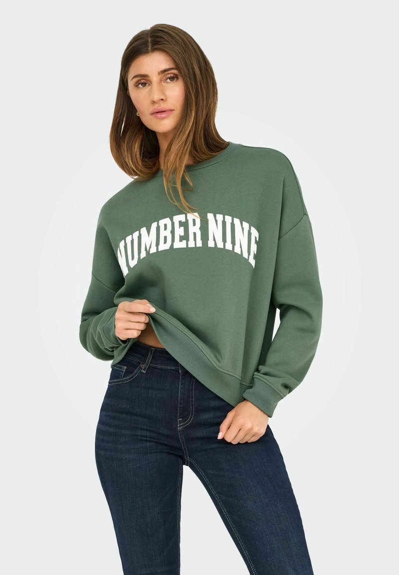 Groene sweatshirt van zacht materiaal met een losse pasvorm, waarop "NUMBER NINE" in vet witte tekst op de voorkant staat. Draagt comfortabel met donkere jeans.