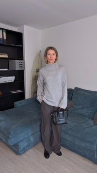 Suéter cinza oversized, calças largas pretas e uma pequena bolsa preta. Texturas suaves com um sofá de veludo azul ao fundo.