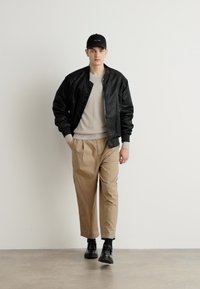 Black bomber jacket, light gray sweater, beige trousers, black shoes, and black cap. Smooth texture, structured design, casual fit.