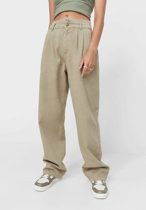 Frau trägt locker sitzende beige Hose, grünes kurzes Tanktop und weiße-grüne Turnschuhe, steht vor einem schlichten hellen Hintergrund.