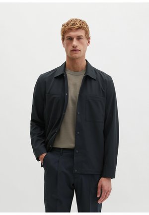 Homme portant une veste noire boutonnée par-dessus un pull gris avec un pantalon foncé, debout devant un fond blanc uni.