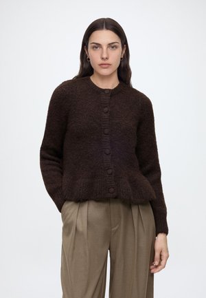 Femme aux longs cheveux foncés portant un cardigan marron foncé duveteux boutonné et un pantalon large plissé beige, debout devant un fond uni.