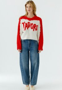 Rode en witte pluizige trui met "J'ADORE" in vetgedrukte letters, gecombineerd met blauw denim wijde pijpen jeans met hoge taille en crèmekleurige schoenen.