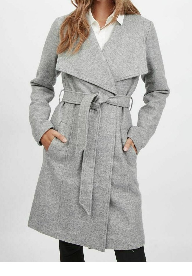 Femme portant un manteau en laine gris clair ceinturé, longueur genoux, avec de larges revers, mains dans les poches, et une chemise blanche en dessous.