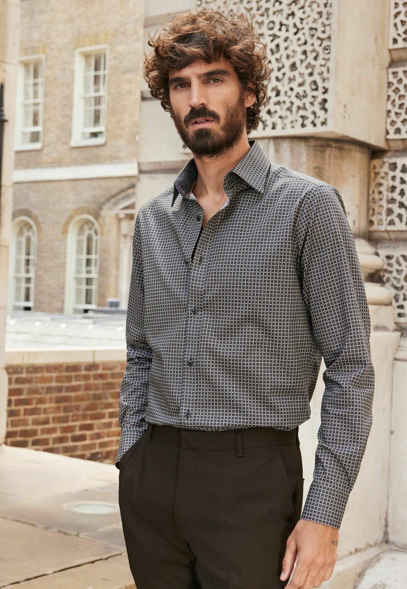 Homme avec des cheveux bouclés et une barbe portant une chemise à motifs et un pantalon sombre, se tenant à l'extérieur près d'un mur en pierre orné.