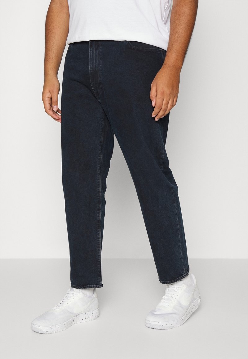 Homme portant un jean bleu foncé, des baskets blanches et un t-shirt blanc, debout devant un fond clair uni.