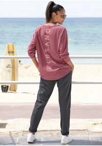 Langärmliges, pinkes Oberteil mit dem "ELBSAND"-Logo auf dem Rücken. Kombiniert mit dunkelgrauen Hosen und weißen Turnschuhen, steht am Strand.