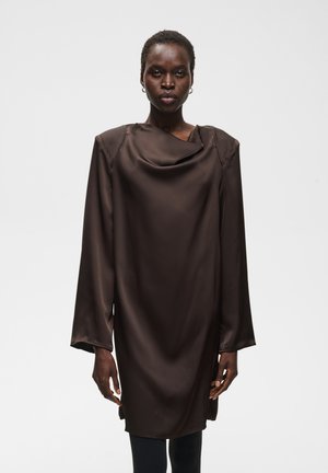 Femme portant une robe en satin marron foncé à manches longues avec un décolleté drapé sur un fond gris clair uni.