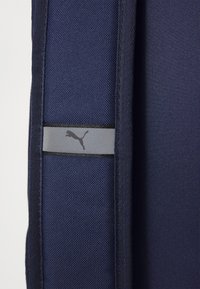 Marineblauw stof met een gestructureerde afwerking, met een horizontale grijze band en een zwart Puma-logo. Het ontwerp is eenvoudig en modern.