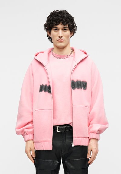 Jeune homme aux cheveux bouclés portant un sweat à capuche rose et un sweat-shirt avec un texte noir en peinture aérosol, une ceinture noire et un pantalon en cuir noir sur fond blanc.