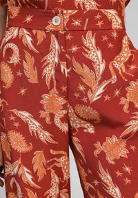 Tissu couleur rouille avec des motifs floraux et de plumes blancs et orange, doté d'une fermeture à bouton et de détails de ceinture.