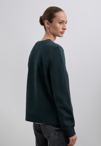 Mujer con el cabello recogido en un moño, vestida con una camiseta de manga larga verde oscuro y jeans negros, girada ligeramente hacia la derecha frente a un fondo simple.