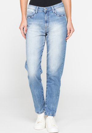 Donna con jeans dritti sbiaditi azzurri chiaro, top nero e sneakers bianche, in piedi davanti a uno sfondo bianco semplice.