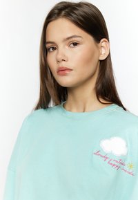 Lichtblauwe sweatshirt van katoen. Kenmerkt zich door een reflecterende borduring van een wolk en de zon aan de linkerkant. Heeft een ronde hals en een comfortabele pasvorm.