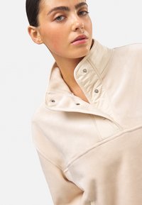 Suéter beige texturizado con cuello alto, que presenta botones a presión y costuras decorativas. Tela suave y corte holgado.