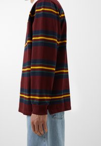 Langärmliges burgunderfarbenes Shirt mit horizontalen Streifen in Gelb, Grün und Dunkelblau. Weicher Stoff, lockere Passform mit seitlichen Schlitzen am Saum.