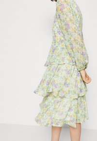 Květinové šaty s vrstvenými cípy v pastelově zelené, žluté a fialové barvě. Lehce průsvitná tkanina s dlouhými rukávy a volánkovými okraji.