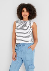 Woman with curly hair wearing a white sleeveless striped top and light blue cargo pants with one hand in pocket against a plain background.