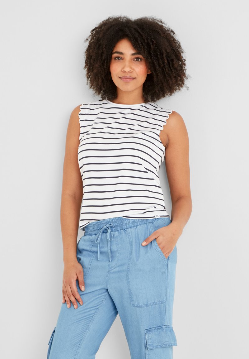 Woman with curly hair wearing a white sleeveless striped top and light blue cargo pants with one hand in pocket against a plain background.