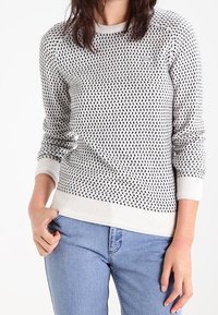 Stickad tröja i vitt med svart mönster, med ribbade ärmslut och nederkant, samt rundad halsringning. Matchad med blå jeans.