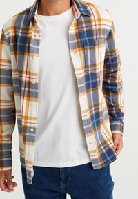 Homme portant un t-shirt blanc à col rond sous une chemise à manches longues à carreaux bleus, jaunes et blancs, non boutonnée, avec un jean bleu.