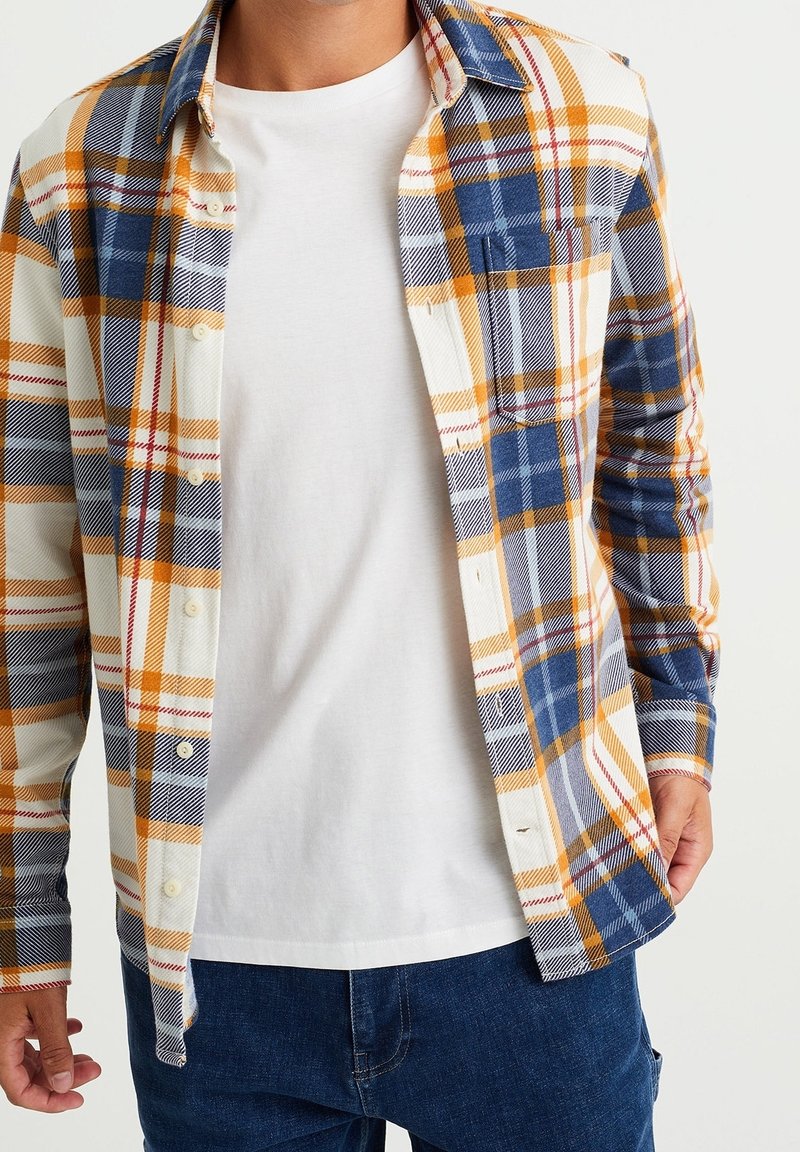 Homme portant un t-shirt blanc à col rond sous une chemise à manches longues à carreaux bleus, jaunes et blancs, non boutonnée, avec un jean bleu.