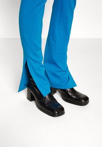 Pantalons ajustés bleus avec des fentes latérales associés à des bottes noires à talons épais en matériau brillant. Lignes épurées, aucun motif visible.