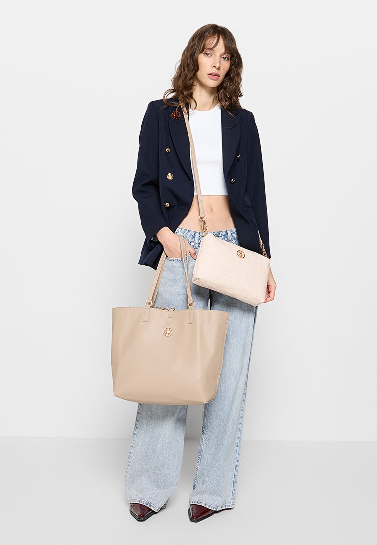 Sac fourre-tout en cuir beige avec deux poignées, logo doré, accompagné d'une petite pochette beige. Le modèle porte un blazer bleu marine et un jean large.