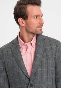 Blazer grigio a houndstooth con quadri rosa, rever a punta e chiusura con un solo bottone, indossato sopra una camicia button-up rosa chiaro.