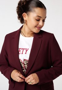 Bordeaux blazer met een slank ontwerp, gecombineerd met een witte T-shirt met graphic in luipaardprint. Gouden sieradenaccenten op de handen.