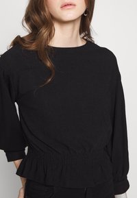 Blusa nera realizzata in tessuto testurizzato, con scollatura rotonda, vita arricciata e maniche ampie, leggermente a sbuffo.