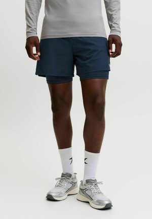 Homme portant un short de sport bleu marine superposé, un t-shirt à manches longues gris clair, des chaussettes blanches montantes et des chaussures de course grises, debout sur fond blanc.