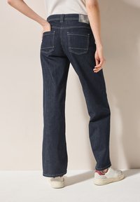 Jean flare en denim bleu foncé avec poches arrière et couture contrastante. Design à taille haute, tissu légèrement texturé, associé à des baskets blanches.