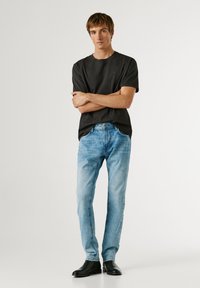 Mladi moški stoji z rokami prekrižanimi, oblečen v črno majico, svetlo modre kavbojke in črne čevlje na enobarvnem svetlem ozadju.