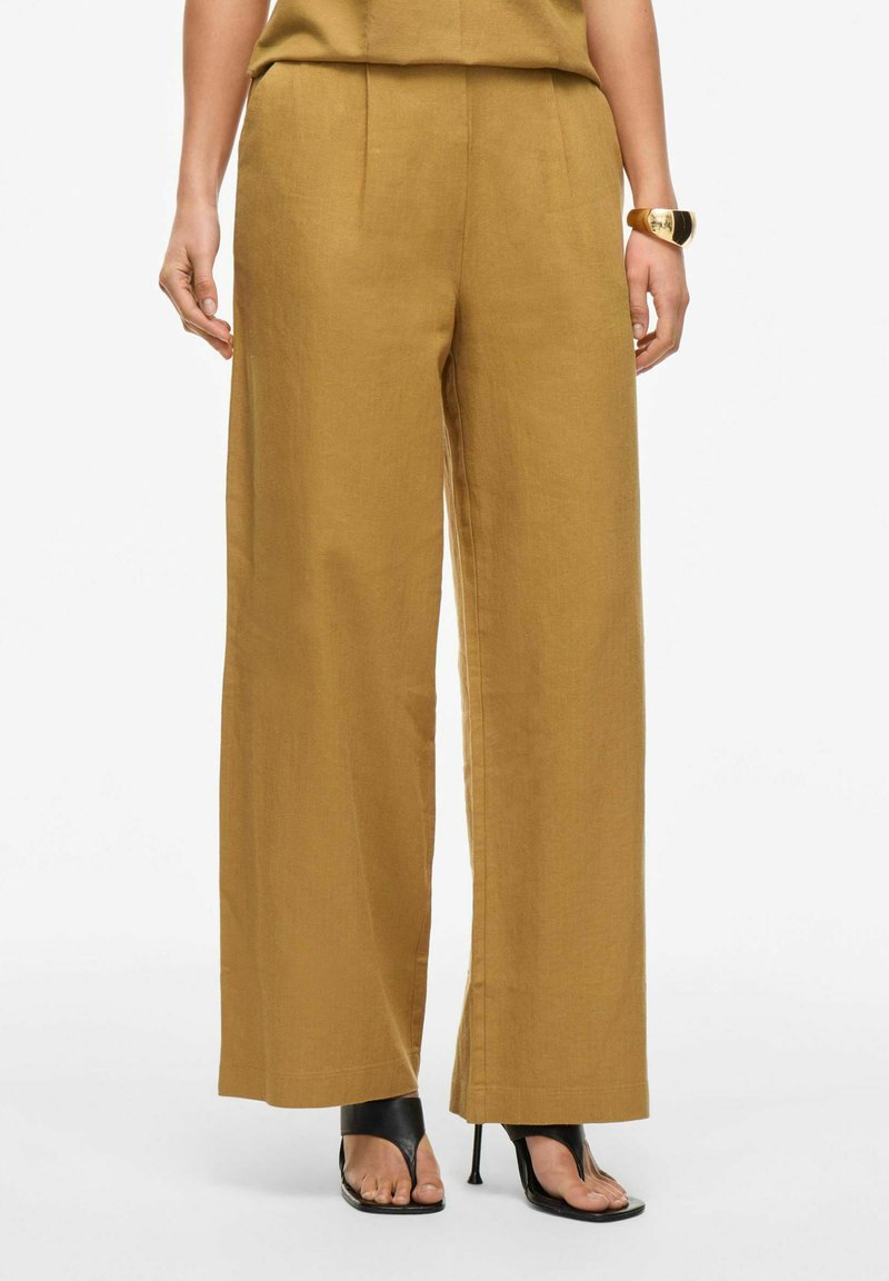 Pantalones de pierna ancha en color mostaza, hechos de una tela texturizada. Presentan una cintura alta y suaves pliegues frontales, combinados con sandalias de tacón negras.