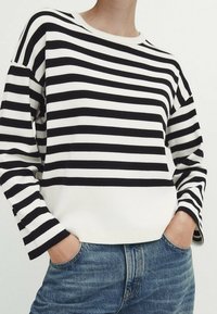 Henkilö, jolla on yllään musta-valkoinen vaakasuora raidallinen pitkähihaainen paita, joka on hieman sisätetty sinisiin farkkuihin ja kädet taskuissa.