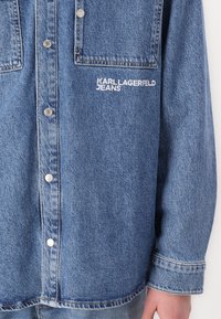 Šviesiai mėlyna džinsinė marškiniai su spaudimo mygtukais, dviem priekinių kišenėmis ir išsiuvinėtu "KARL LAGERFELD JEANS" logotipu ant krūtinės.