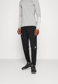 Camisa de manga larga gris claro con rayas blancas en las mangas, combinada con pantalones negros que tienen un logo blanco y zapatillas deportivas negras.