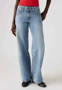 Lichtblauwe wijduitlopende denim jeans met een hoge taille, metalen knoopsluiting en subtiele vervaging. Gecombineerd met zwarte platte schoenen.