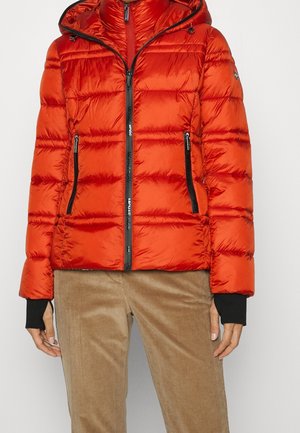 Veste matelassée orange vif avec poches zippées noires et capuche, portée avec un pantalon en velours côtelé beige et des poignets de manches noirs visibles.
