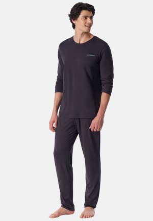 Homme portant un pyjama sombre à manches longues et pantalon, debout pieds nus et regardant sur le côté sur un fond uni.