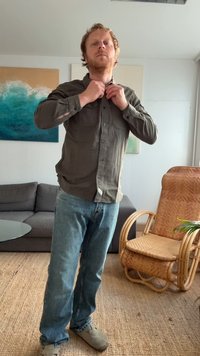 Camisa verde-oliva de botão, jeans de ganga azul-claro, sapatos bege. Pessoa a ajustar a gola da camisa, de pé numa sala com um sofá.