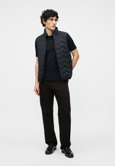 Νεαρός άνδρας με σγουρά μαλλιά, φοράει μαύρο καπιτονέ γιλέκο πάνω από μαύρο πόλο, σκούρο παντελόνι και μαύρα επίσημα παπούτσια, στέκεται με τα χέρια πίσω από την πλάτη.