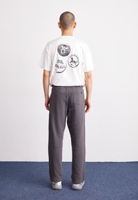 Weißes Baumwoll-T-Shirt mit grafischen Drucken auf dem Rücken, kombiniert mit grauen Sweatpants mit einer Gesäßtasche. Modell steht vor einem schlichten Hintergrund.