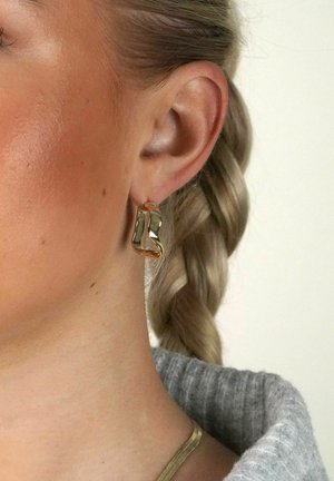Goldene Ohrringe mit einem gewellten, abstrakten Design. Das Licht spiegelt sich auf den glatten Oberflächen und verstärkt den metallischen Glanz und die Konturen.